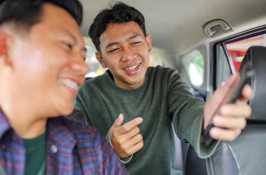 Two men smiling in the back of a car as the younger one verifies ride data on his phone.