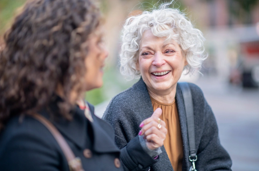 Two women chatting and smiling on a city sidewalk before NEMT ride, one with silver curly hair.