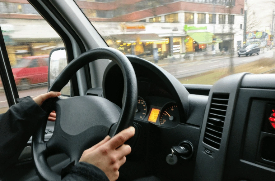 A first-person point-of-view shot from the driver's seat of a commercial van. A hand is on the steering wheel, and the dashboard is visible, showing gauges and an ignition key. Through the windshield and side windows, motion-blurred city streets and buildings are visible.